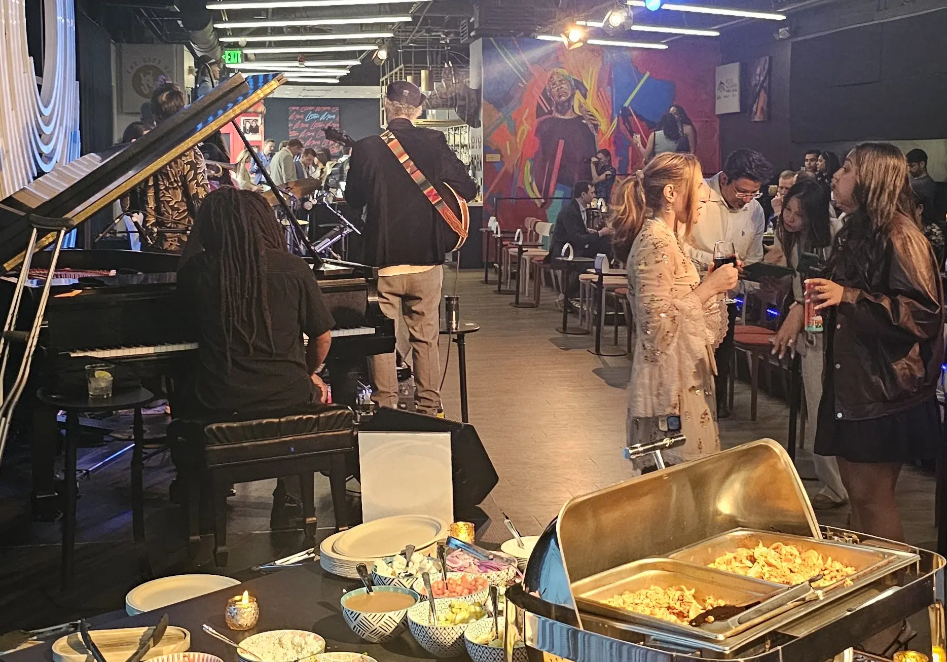 Chicken tinga tostada food station in the foreground with musicians on stage on the right and people talking on the left