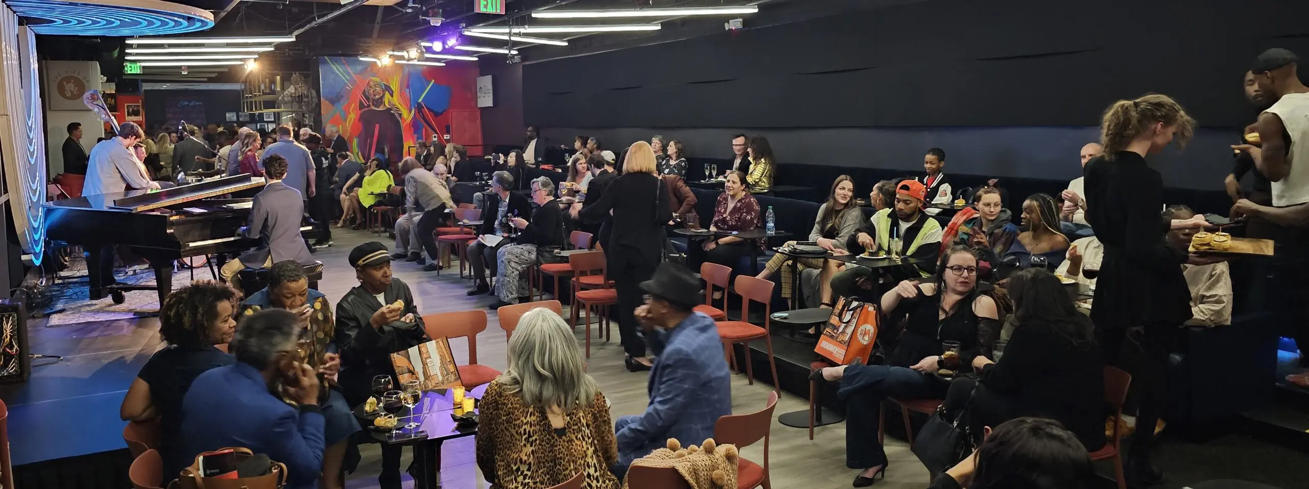 People eating and drinking during a party at Dazzle in Denver, CO. There's a band on the stage on the left and a server with a tray of sliders on the right.